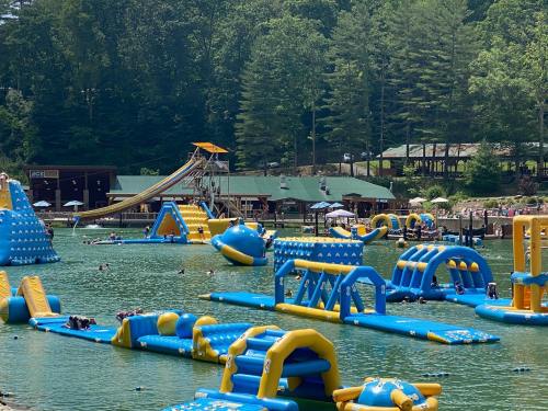 A vibrant water park with inflatable structures on a lake, surrounded by trees and people enjoying the sun.