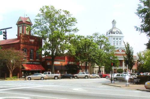 Historic downtown scene featuring a red brick building and a clock tower, with trees lining the street and parked cars.