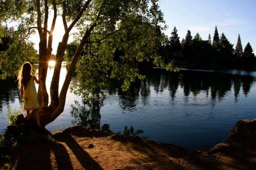 A girl in a yellow dress stands by a tree near a calm river at sunset, with reflections on the water.