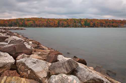 A rocky shoreline with calm water and autumn-colored trees in the background under a cloudy sky.