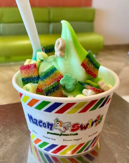 A colorful cup of green frozen yogurt topped with rainbow candy and cereal, served with a white spoon.