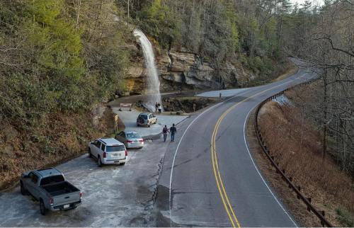 A scenic view of a waterfall beside a winding road, with parked cars and people near the water's edge.