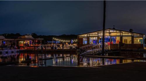 A waterfront restaurant at night, illuminated with colorful lights, reflecting on calm water.
