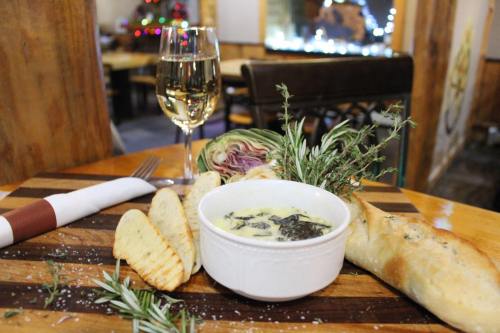 A wooden table with a bowl of creamy dip, bread slices, herbs, and a glass of white wine in a cozy restaurant setting.