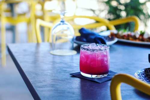 A vibrant pink cocktail with ice, served in a glass on a table, with yellow chairs and a blurred background.