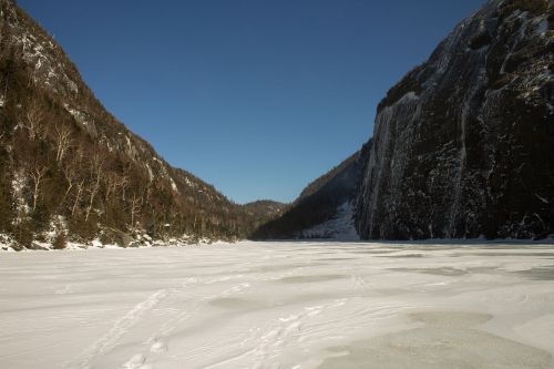 A frozen landscape with snow-covered ground, flanked by steep, rocky cliffs under a clear blue sky.