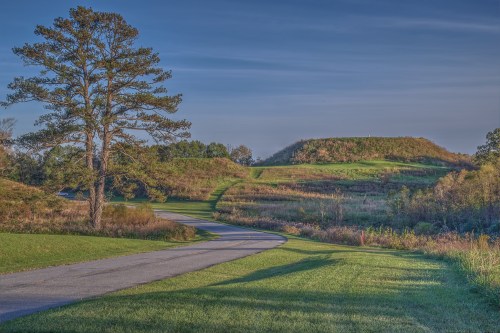 A winding road leads through grassy fields, with a large mound and trees under a clear blue sky.