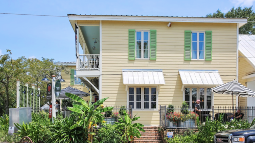 A charming yellow house with green shutters, featuring outdoor seating and lush greenery in a sunny setting.