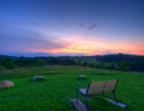A serene landscape at sunset, featuring a bench overlooking a grassy field and distant hills under a colorful sky.