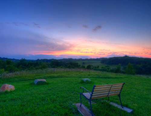 A serene landscape at sunset, featuring a bench overlooking a grassy field and distant hills under a colorful sky.