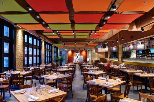 Modern restaurant interior with wooden tables, colorful ceiling panels, and a large mural of a man on the wall.