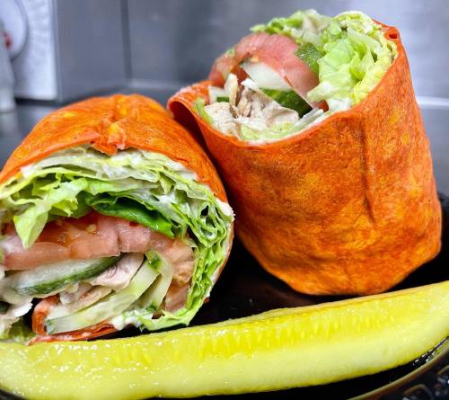 Two colorful wraps filled with lettuce, tomatoes, cucumbers, and chicken, served with a pickle on the side.