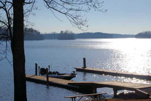 A serene lake scene with a boat docked at a wooden pier, surrounded by trees and shimmering water under a clear sky.