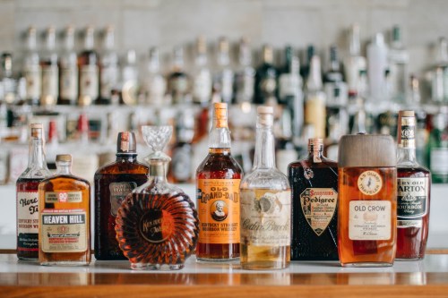 A variety of whiskey and liquor bottles displayed on a bar counter, with a blurred background of more bottles.