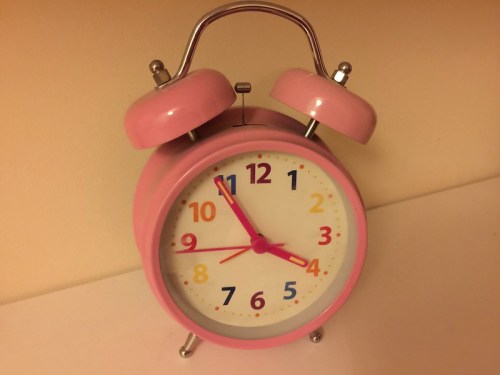 A pink alarm clock with colorful numbers and two bells on top, set against a light background.
