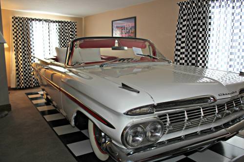 A classic white convertible car parked in a retro-themed room with checkered curtains and flooring.