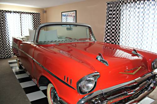 A bright red vintage convertible car parked indoors, with checkered curtains and flooring in the background.