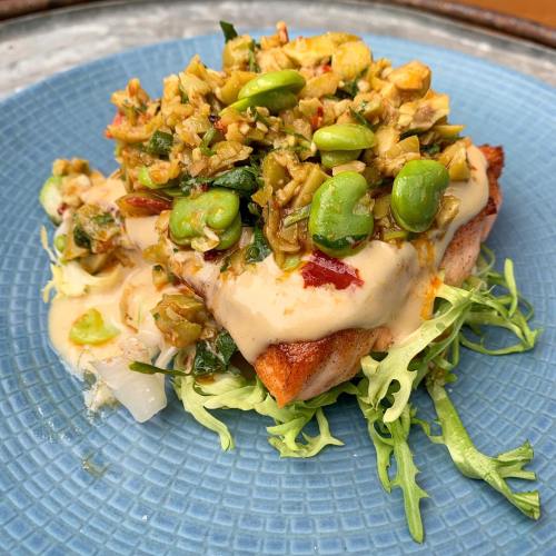 A colorful dish featuring a piece of fish topped with a vibrant vegetable mix and creamy sauce, served on a blue plate.