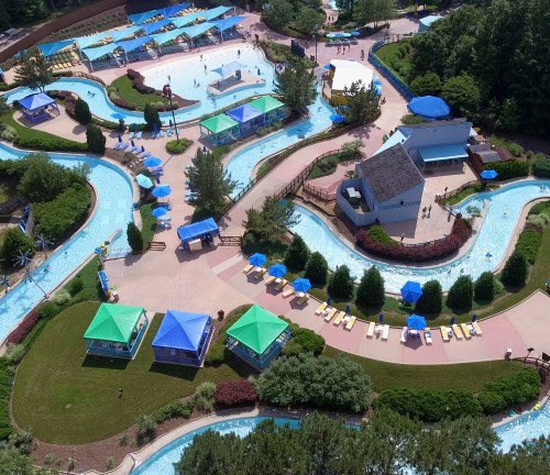Aerial view of a water park featuring winding lazy rivers, cabanas, and sun loungers surrounded by greenery.