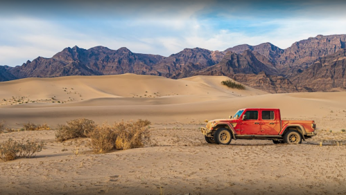 A red truck parked on sandy dunes with mountains in the background under a clear sky.