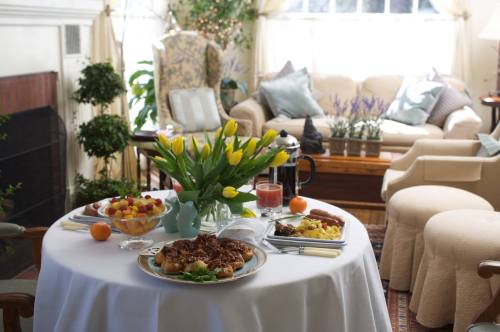 A beautifully set table with a floral centerpiece, featuring a variety of dishes and a cozy living room in the background.
