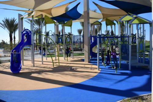 Colorful playground with slides and climbing structures, surrounded by palm trees and a sunny blue sky.