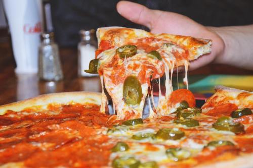 A hand pulls a slice of pepperoni pizza topped with jalapeños, cheese stretching from the pie.