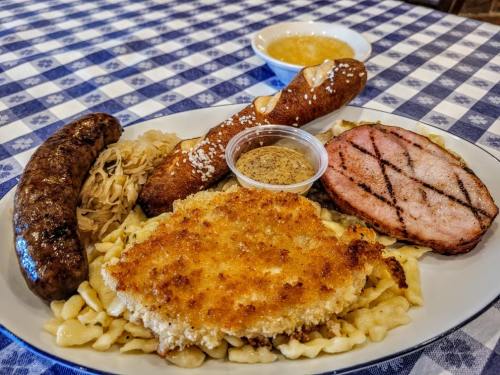A plate of German cuisine featuring schnitzel, sausage, ham, pretzel, sauerkraut, and mustard on a checkered tablecloth.