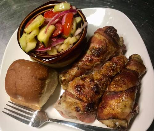 A plate with grilled chicken legs wrapped in bacon, a roll, and a side of salad with cucumbers and peppers.