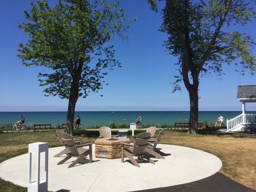 A serene lakeside scene with chairs around a fire pit, trees, and cyclists in the background under a clear blue sky.
