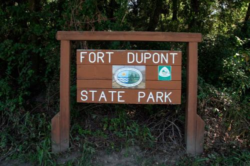 Wooden sign for Fort Dupont State Park, featuring park logos and surrounded by greenery.