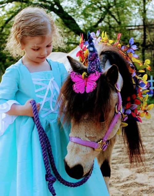 A young girl in a blue dress gently holds a unicorn-themed pony adorned with flowers and butterflies.