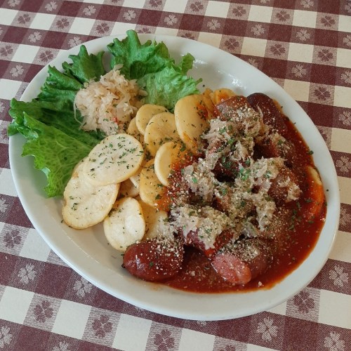 A plate of sausage in sauce, sliced potatoes, lettuce, and sauerkraut on a checkered tablecloth.