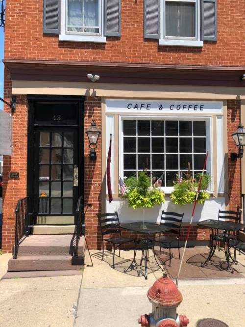 A charming café with outdoor seating, featuring a brick facade and large windows, adorned with plants and flags.