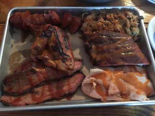 A metal tray filled with various smoked meats, including brisket, chicken, sausage, and pulled pork, drizzled with sauce.