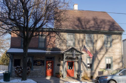 Historic stone building with a sloped roof, red doors, and an American flag, surrounded by snow and trees.