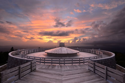 A circular wooden observation deck at sunset, with vibrant clouds and a dramatic sky in the background.