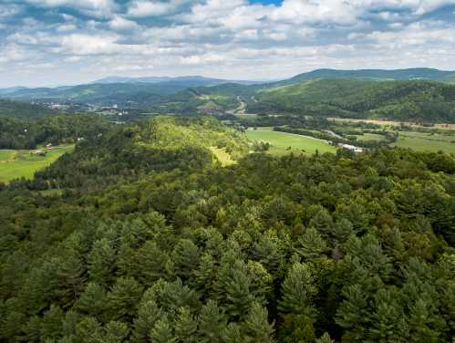 Aerial view of lush green hills and forests under a partly cloudy sky, with valleys and distant mountains visible.