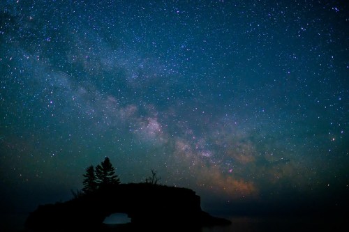 A starry night sky filled with the Milky Way over a dark silhouette of a rocky island and trees.