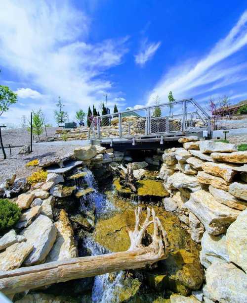 A serene landscape featuring a small waterfall flowing over rocks, with a bridge and blue sky in the background.