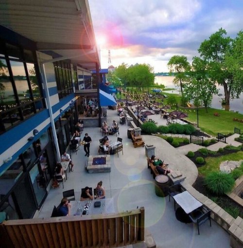 A lively outdoor dining area by a lake, with people enjoying food and drinks as the sun sets in the background.