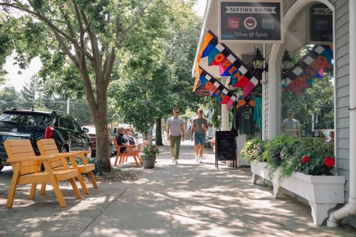 A sunny street scene with people walking past shops, colorful flags, and outdoor seating. Trees line the sidewalk.
