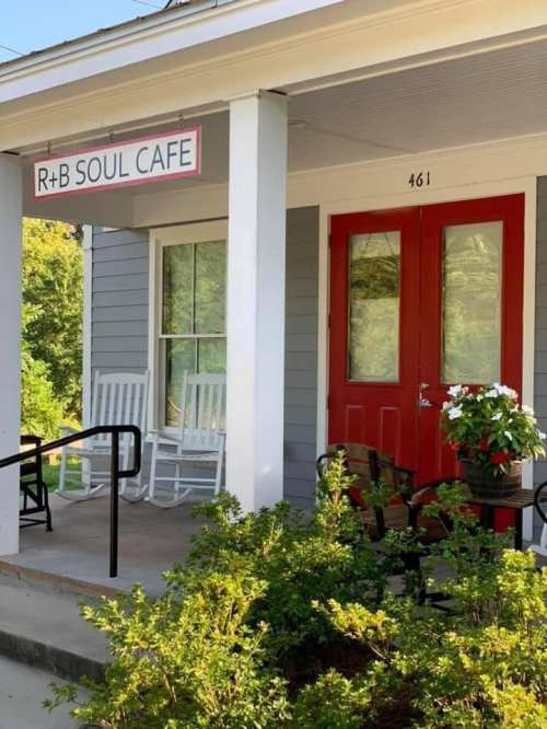 Exterior of R+B Soul Cafe featuring a red door, sign, and seating area surrounded by greenery.