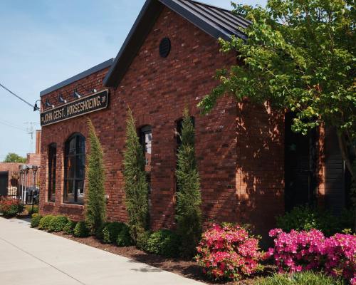 A brick building with a sign reading "John Cest. Horseshoeing," surrounded by greenery and colorful flowers.