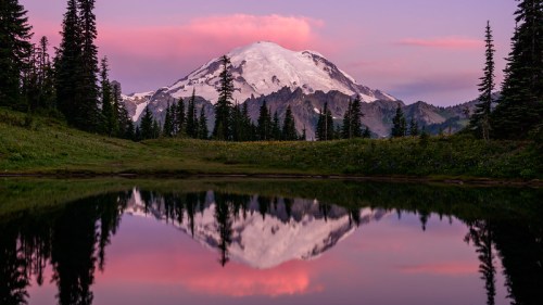 A serene landscape featuring a snow-capped mountain reflecting in a calm lake, surrounded by lush trees at sunset.