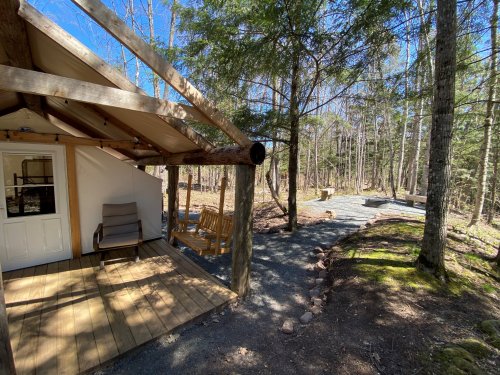 A cozy cabin with a porch in a wooded area, surrounded by trees and a stone pathway leading to a fire pit.