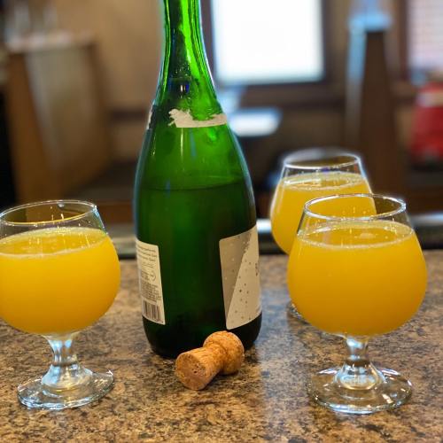 Four glasses of orange mimosa with a green bottle and a cork on a countertop.