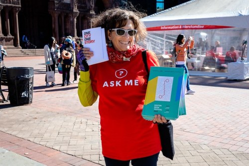 A woman in a red "ASK ME" shirt holds brochures at an outdoor event, smiling in front of a tent.