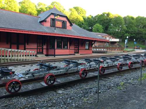 A red train station with a row of rail cars on tracks in the foreground, surrounded by green trees.