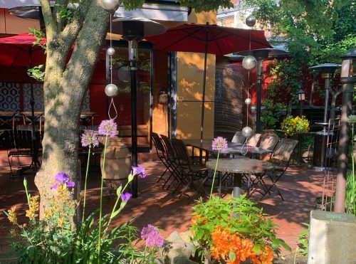 A cozy outdoor dining area with tables, umbrellas, and colorful flowers under a tree.
